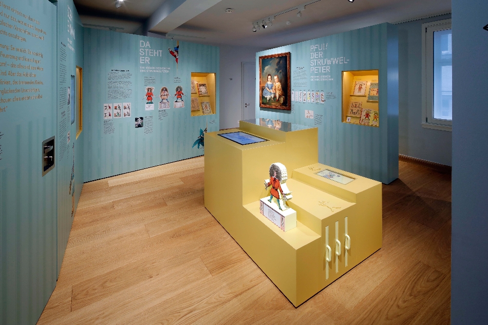 Bild vergrössern: Ausstellung im Struwwelpeter Museum mit hellblauen Wänden und gelbem Erkundungstisch im Vordergrund