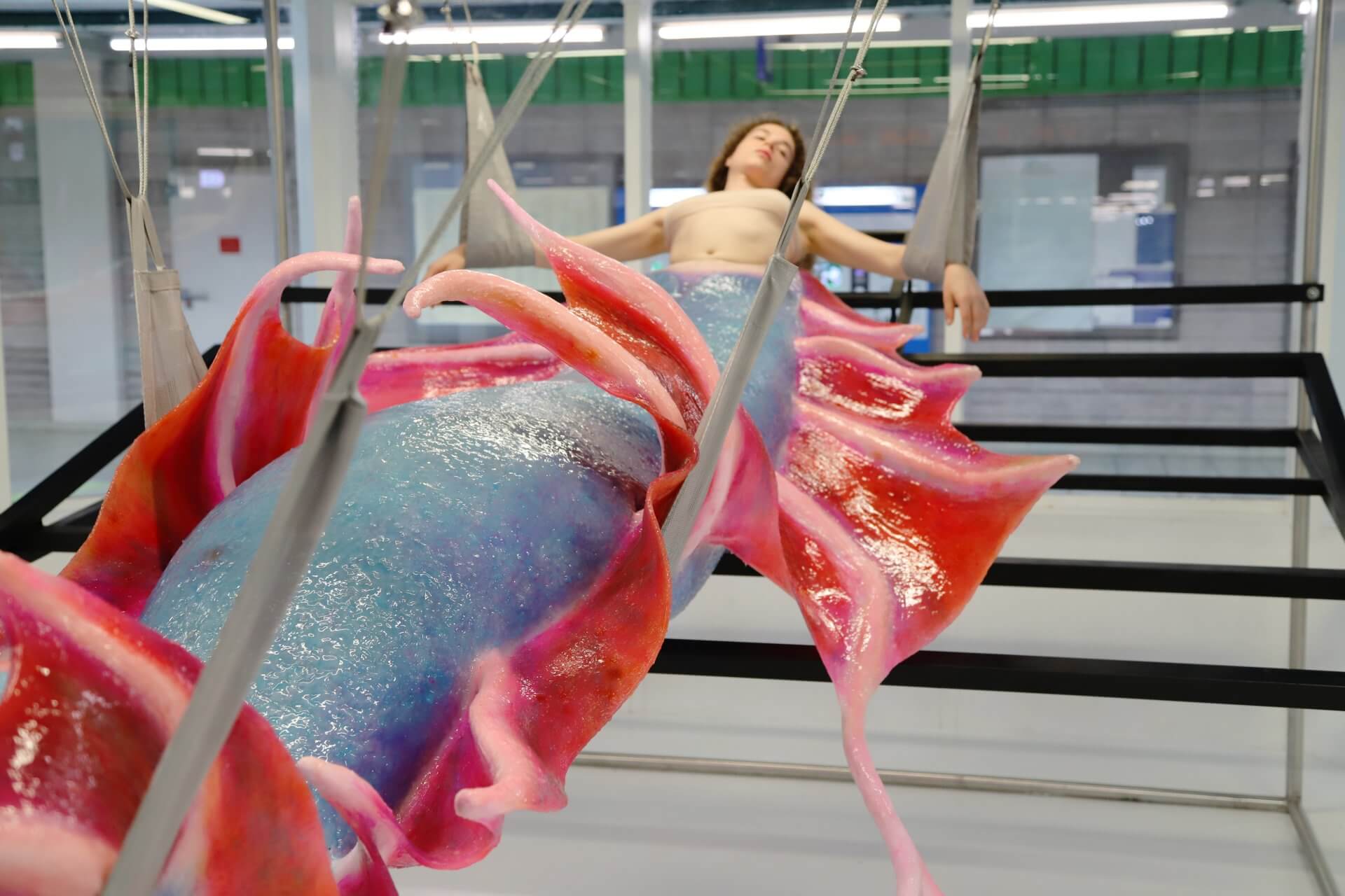 Agnes Questionmark, CHM13hTERT, 2023, performance and installation, 2023 , curated by The Orange Garden and Spazio Serra supported by Kuboraum 2023, Photo: Cristiano Rizzo, ©Agnes Questionmark, Courtesy: the artist Frau hängt in einem Raum mit rot-blauem Fisch-Kostüm am Unterkörper
