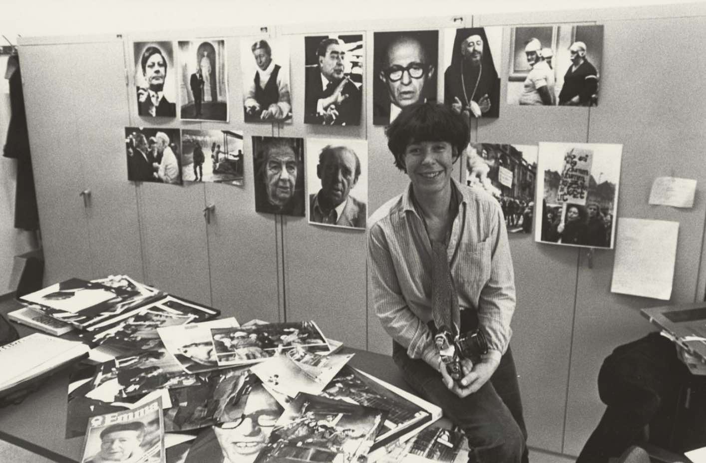 Schwarz-weiß Fotografie, auf der eine Frau mit kurzen dunklen Haaren auf einem Tisch sitzt mit vielen Fotos und Magazinen, im Hintergrund Portäts an der Wand