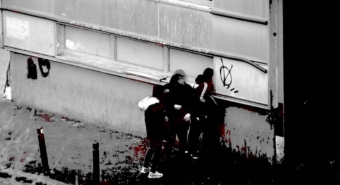 Bild vergrössern: Verschwommenes schwarz-weiß Foto dreier Personen mit Jogginganzügen und Kopfbedeckung von hinten, die durch einen Fensterspalt in ein heruntergekommenes Gebäude blicken