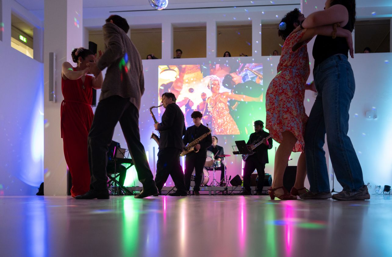 Zwei Paare tanzen in einem wei&szlig;en Raum vor einer Band und einer Projektion an der Wand, durch Durchbr&uuml;che in der Wand schauen von oben Personen zu