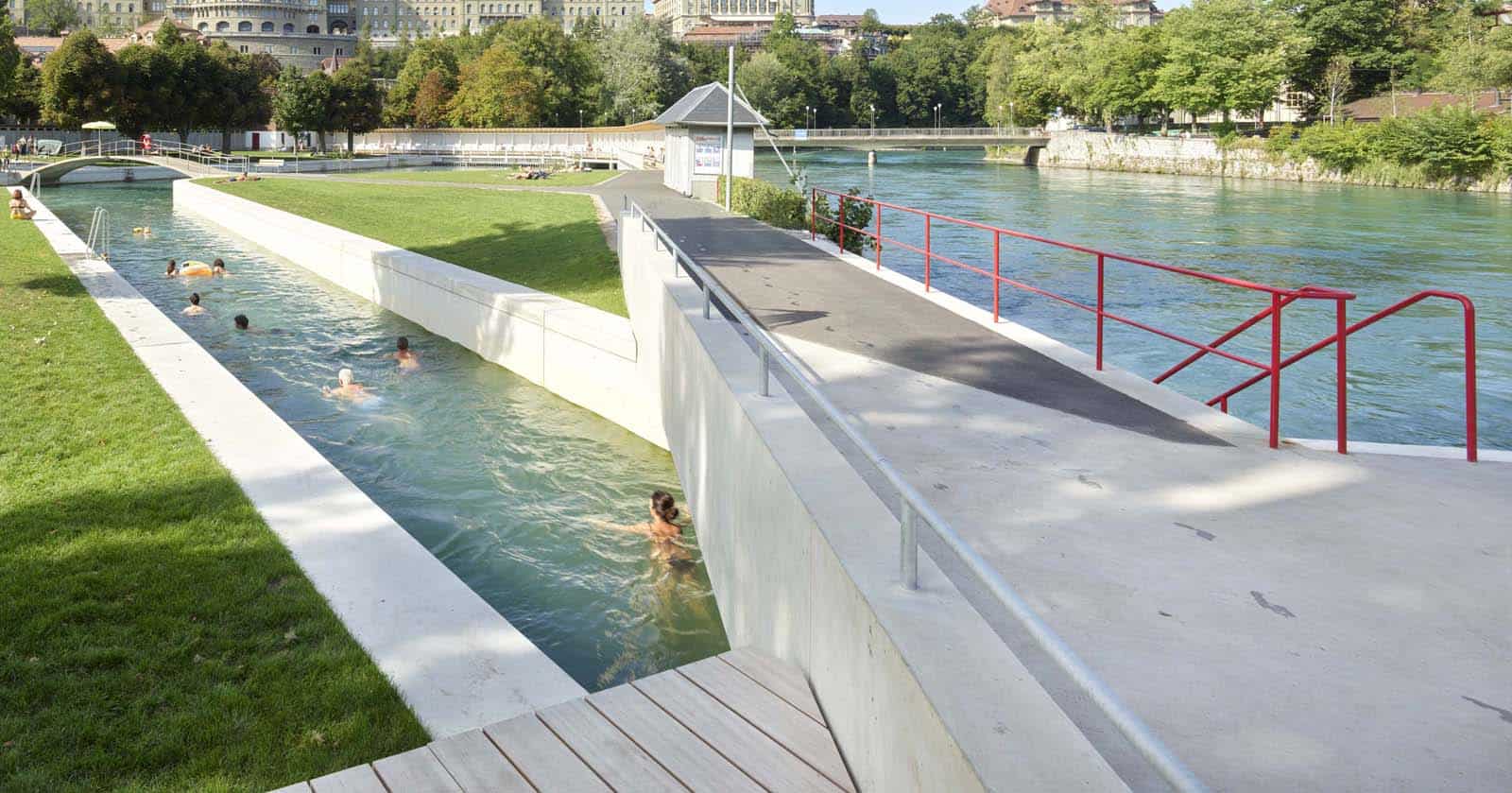 Bild vergrössern: Mehrere Menschen schwimmen in einem Kanal neben einem Fluss in einer Stadt