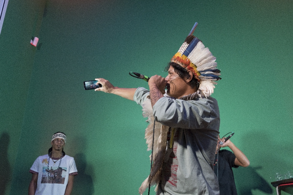 Bild vergrössern: Rapper mit Mikrophon und Handy in der Hand sowie Indianerhäuptlings-Federschmuck auf dem Kopf im Vordergrund rechts und Rapper mit Bandana im Hintergrund links im Weltkulturen Museum