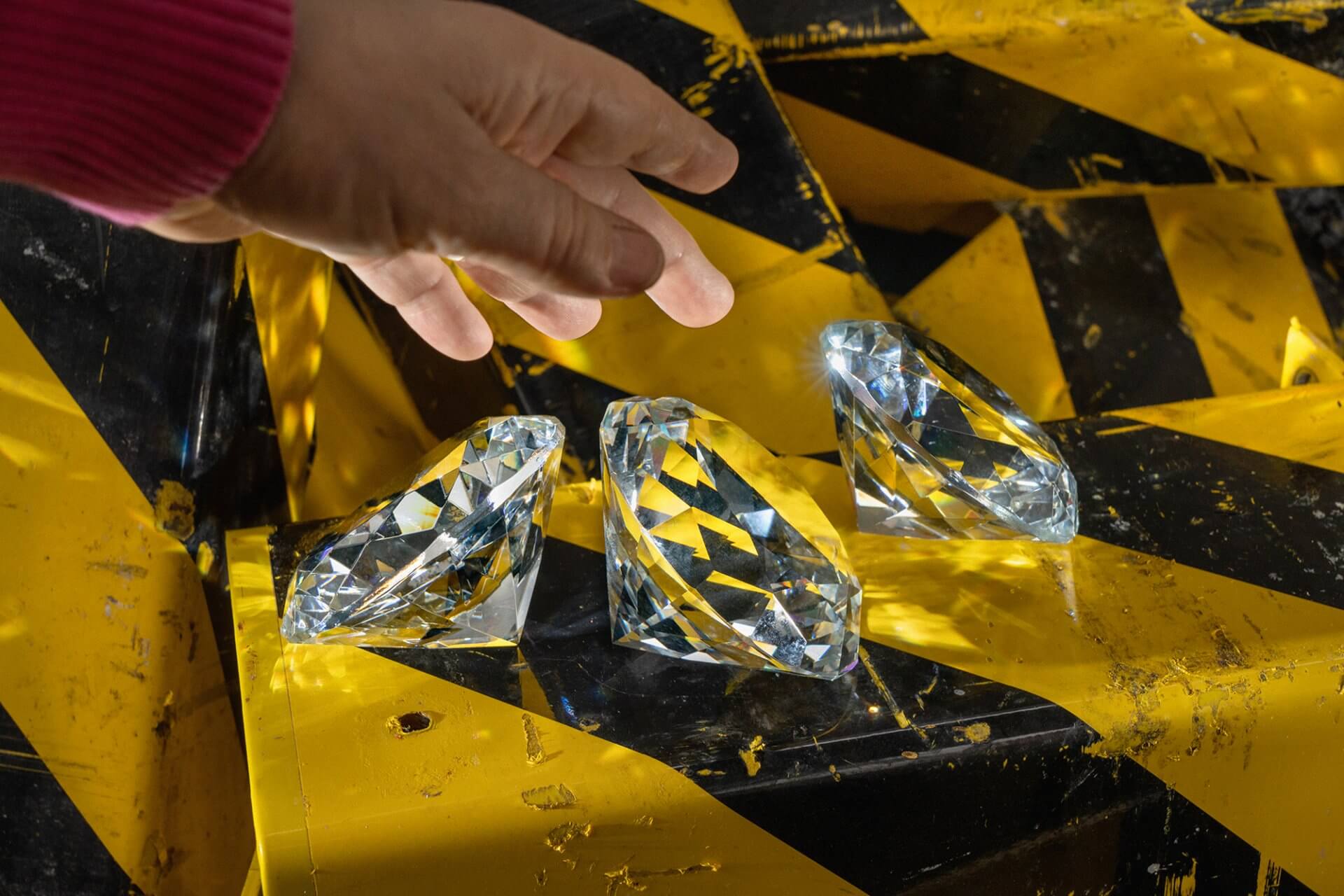 Bild vergrössern: Hand, die nach großen Diamanten greift, die auf einem gelb-schwarzem Untergrund liegen