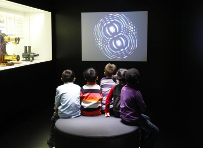 Enlarge image: Kinder sitzen vor Bildschirm und Vitrine in einem abgedunkelten Raum im Deutschen Filmmuseum