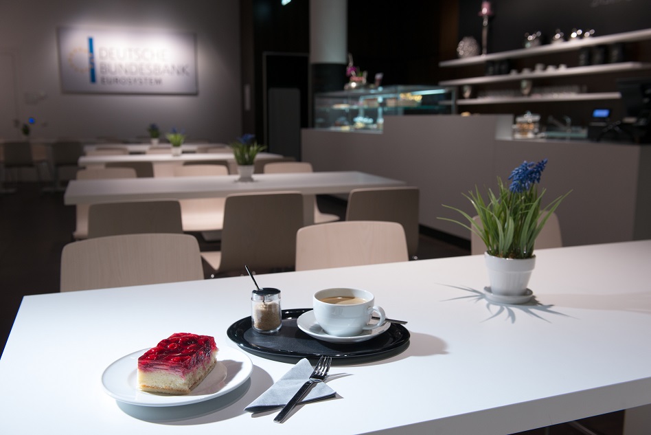 Blick auf einen Tisch mit einem Teller mit einem Stück Erdbeertorte und einer Tasse Kaffe und der Theke der Cafeteria des Geldmuseums im Hintergrund