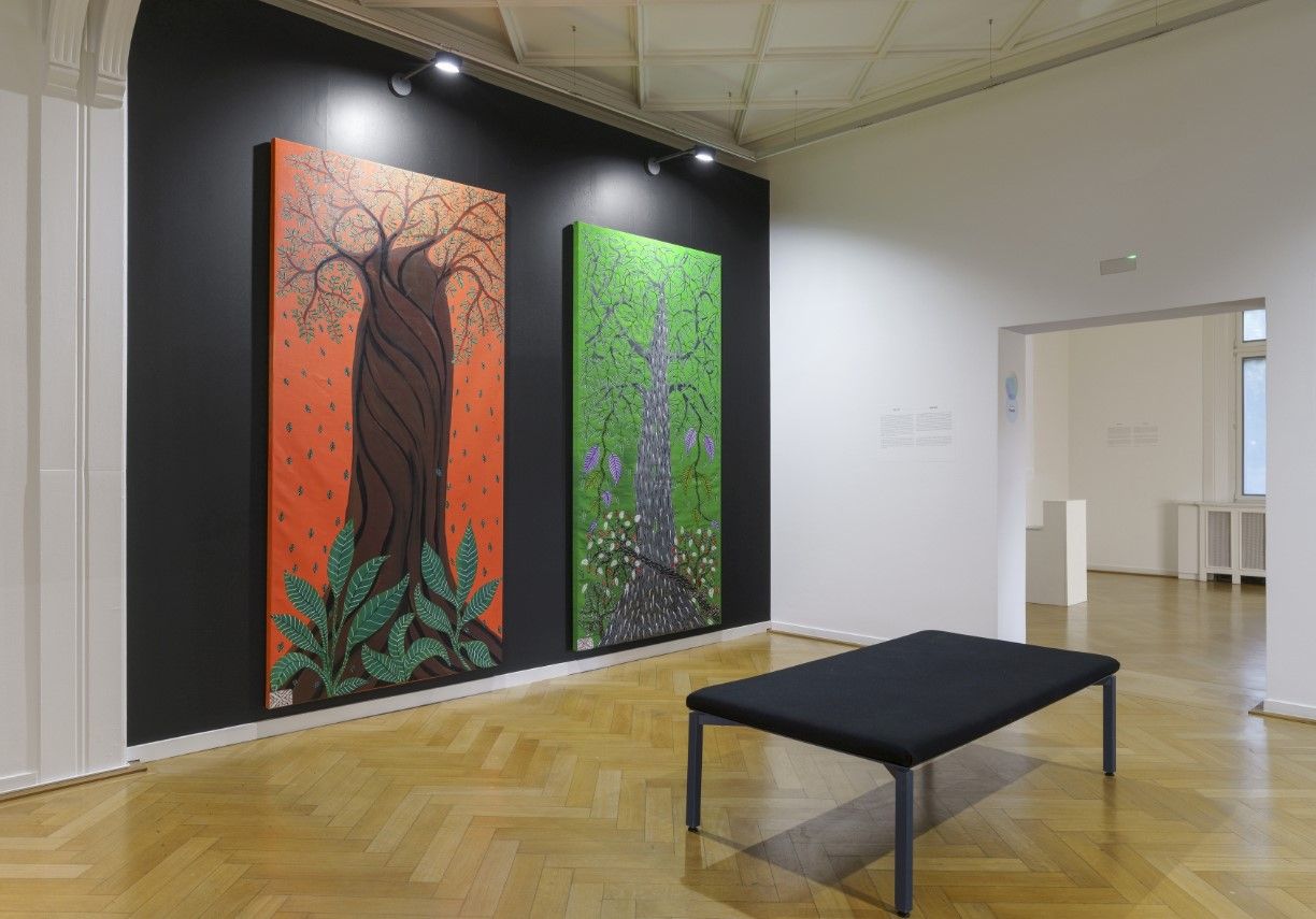 Zwei große Gemälde mit Bäumen darauf hängen links vor einer schwarzen Wand im Ausstellungsraum, das eine Bild hat einen orangenen Hintergrund, das andere einen grünen, rechts im Vordergrund eine schwarze Bank