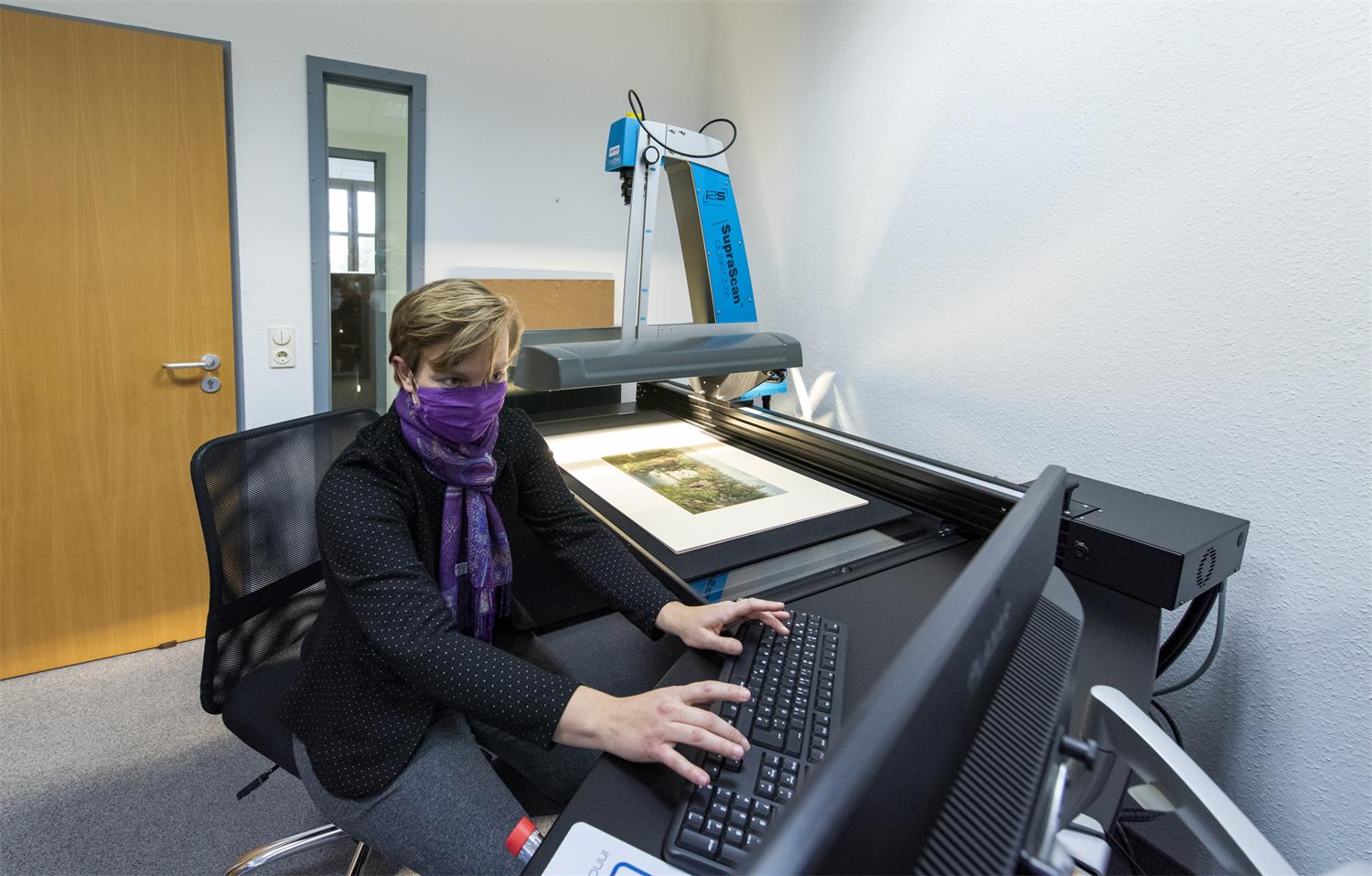 Eine Mitarbeiterin mit Schal vorm Gesicht sitzt vor dem Scanner an einem Bildschirm
