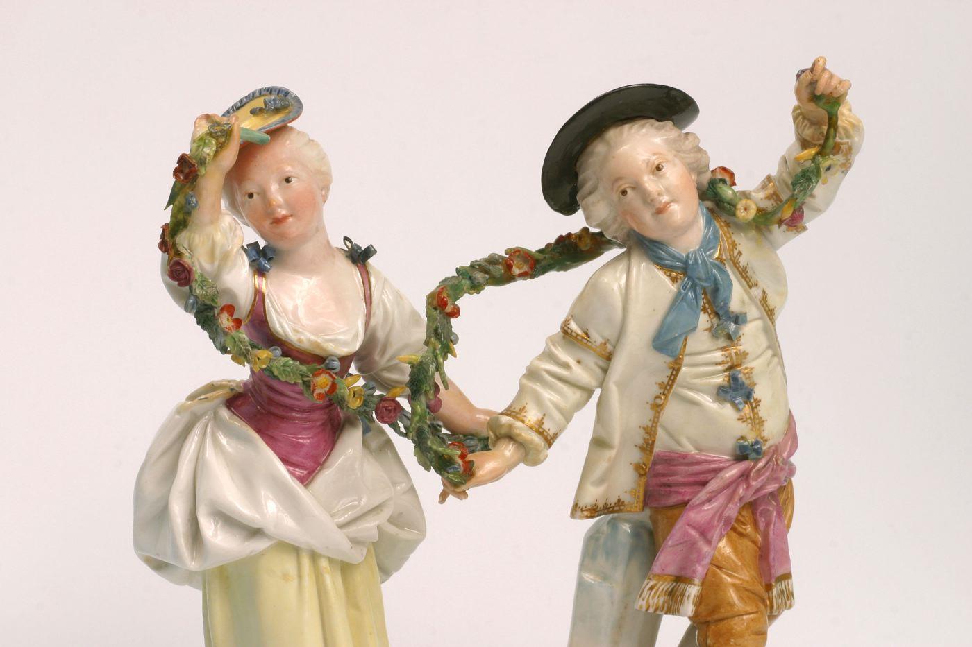 Ein Paar aus Porzellan tanzt miteinander, sie halten eine Girlande in den Händen