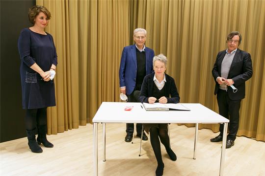 Die Kulturdezernentin unterschreibt ein Dokument, Mirjam Wenzel, Salomon Korn und Marc Grünbaum schauen ihr dabei zu.