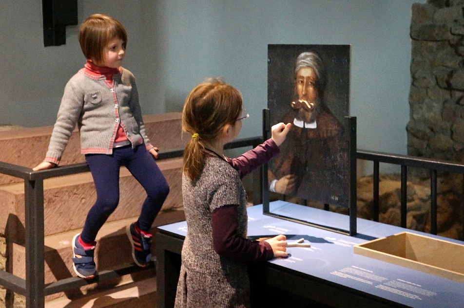 Zwei Mädchen im Museum Judengasse blicken auf eine Nachbildung eines Porträts und eines hält dem Mann auf dem Gemälde einen goldenen Bart vor