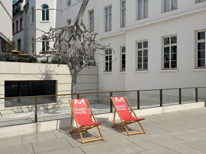 Zwei rote Liegestühle mit der Aufschrift Kunstpause auf einem Platz vor einem Gebäude