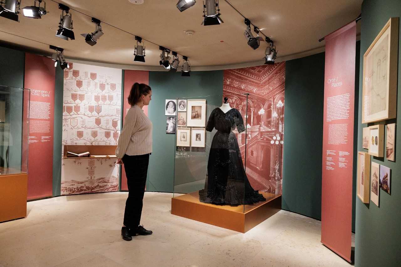 Enlarge image: Eine Frau steht in einem Ausstellungsraum und schaut auf ein schwarzes langes Kleid in einer Vitrine