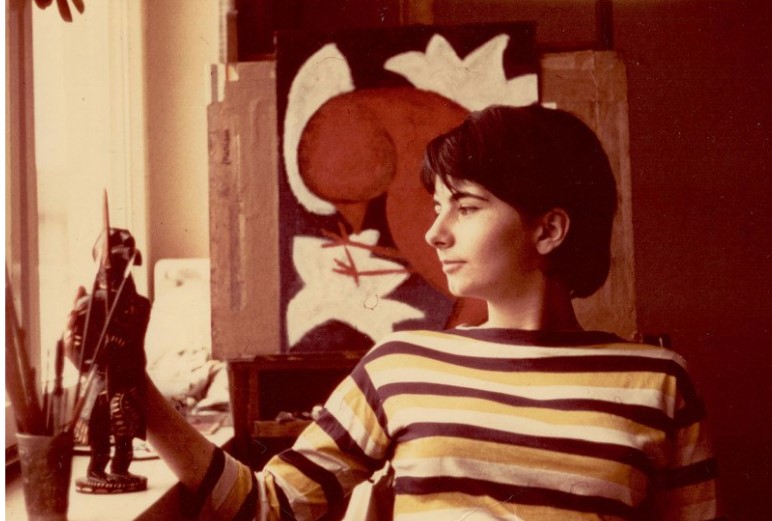 Auf dem sepiafarbenen Foto schaut ein Mädchen auf Pinseln, im Hintergrund ein Gemälde mit einem Vogel