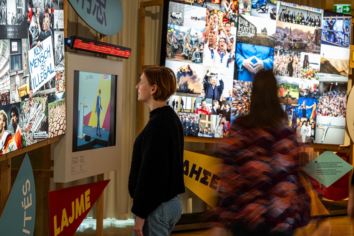 Bild vergrössern: Zwei Personen stehen in Raum mit Bildschirmen und Nachrichtenbilderflut an den W&auml;nden und schauen diese an