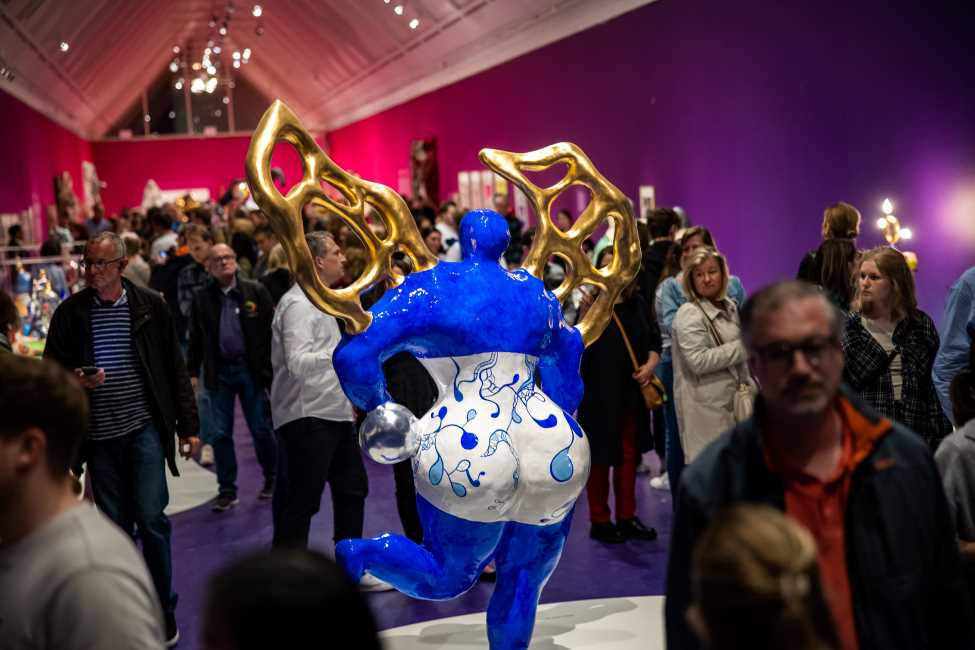 Viele Menschen in einem Ausstellungsraum, in dem in der Mitte eine blau-weiße abstrakte Skulptur einer rundlichen Frau mit goldenen Flügeln steht