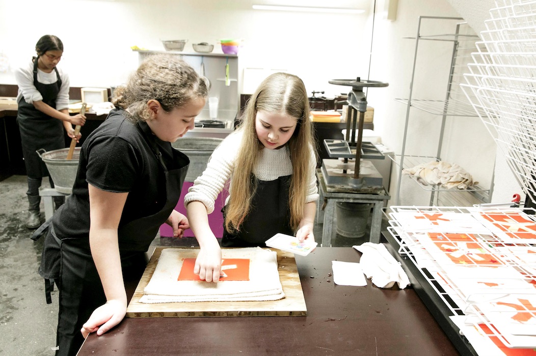 Bild vergrössern: Zwei Mädchen beugen sich über einen Tisch in der Druckwerkstatt, wo sie einen Druck des Logos des Museums herstellen, im Hintergrund rührt ein weiteres Mädchen in einem Zuber