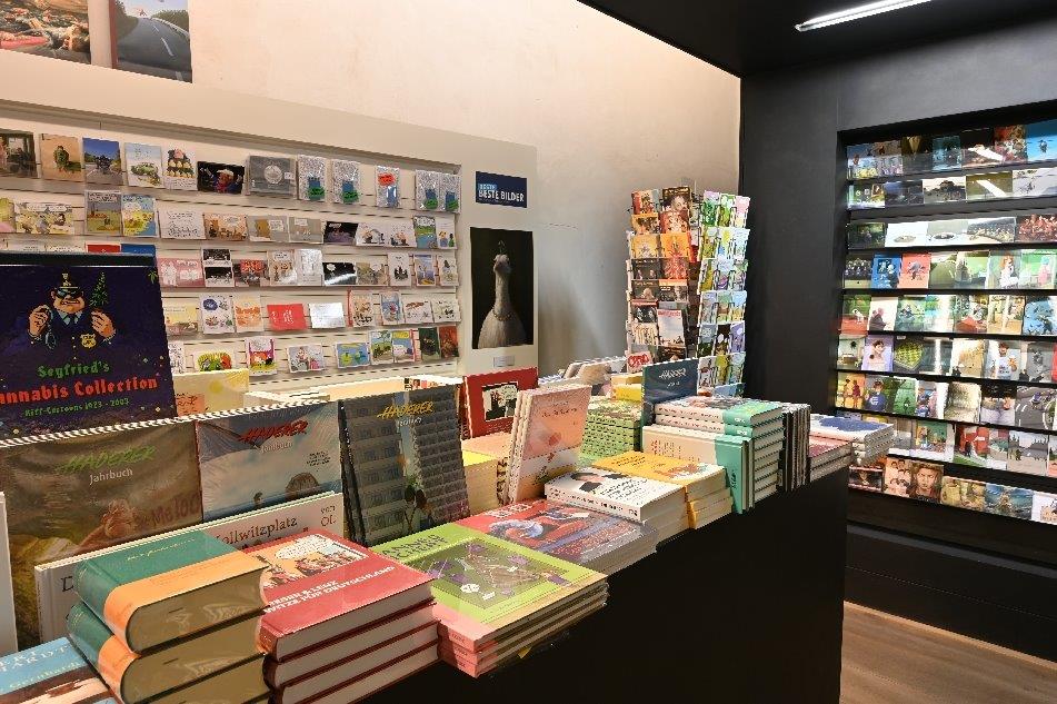 Ansicht des Shops im Caricatura Museum mit gestapelten Büchern im Vordergrund und einem Postkartenregal links hinten und einem Postkartenständer und Regal rechts hinten