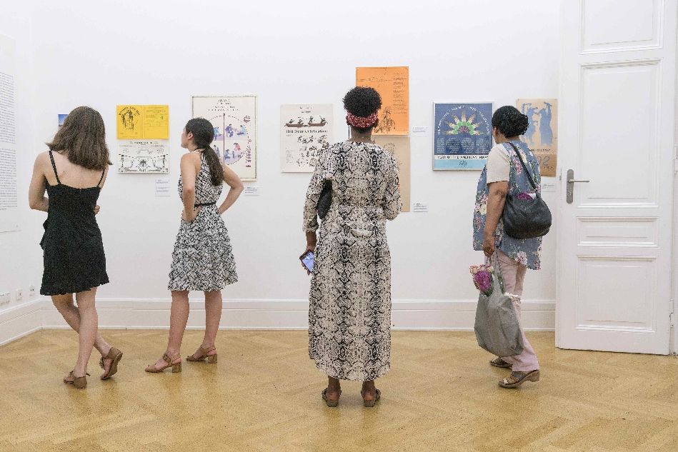 Vier Besucherinnen (von hinten fotografiert) betrachten Plakate und Entwürfe in einer Ausstellung im Weltkulturen Museum