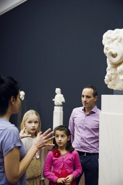 Zwei Kinder mit Vater in der Skulpturenausstellung hören Frau im Vordergrund zu 