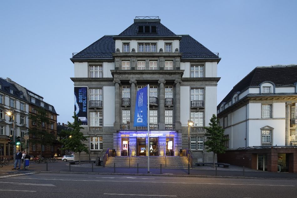 Fassade des Filmmuseum mit Straße im Vordergrund und blau erleuchtetem Eingangsbereich
