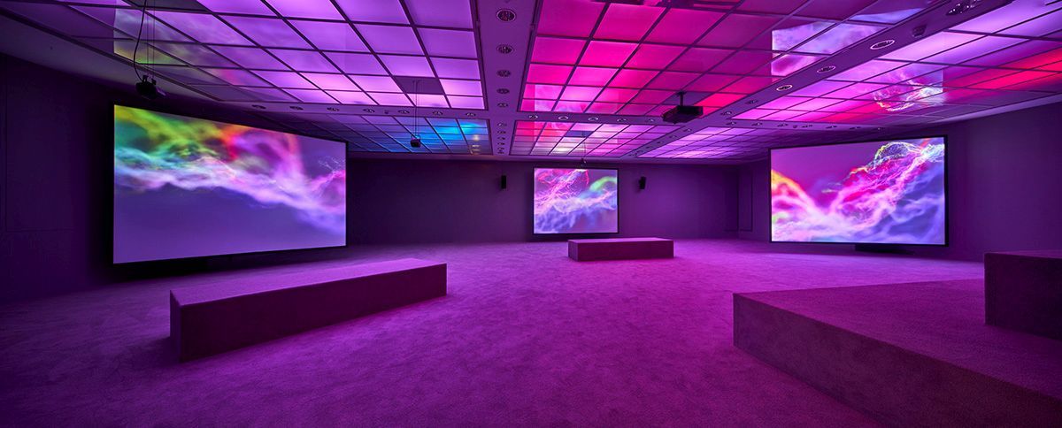 Drei große Screens zeigen bunte Lichtwellen in einem in rosa-lila Licht getauchten Ausstellungsraum 