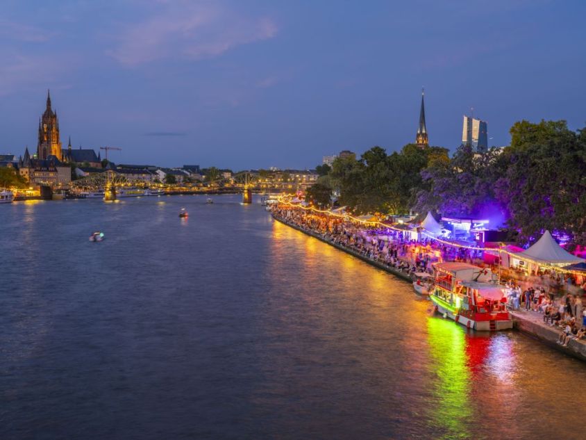 Erleuchtetes Ufer eines Flusses mit Buden und vielen Menschen von Weitem mit Brücke, Hochhaus und Kirchen im Hintergrund