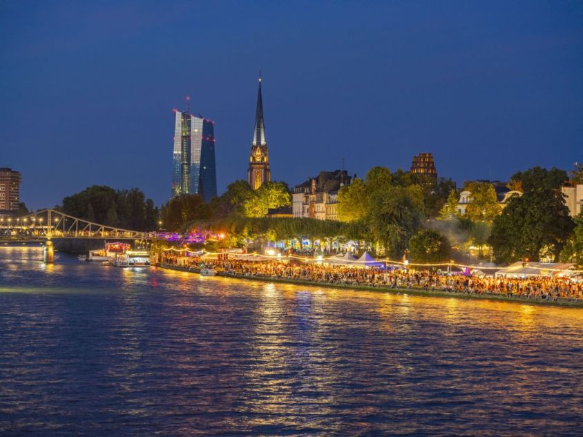 Erleuchtetes Ufer eines Flusses mit Buden und vielen Menschen von Weitem mit Brücke, Hochhaus und Kirche im Hintergrund