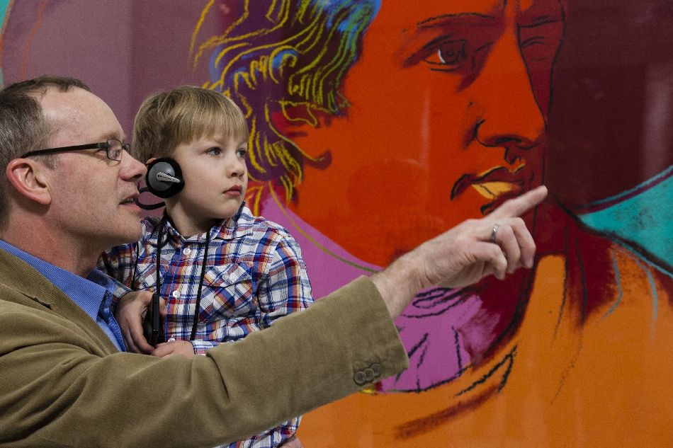 Vater mit Sohn auf dem Arm links zeigt auf etwas außerhalb des Fotos, im Hintergrund die Grafik "Johann Wolfgang von Goethe" von Andy Warhol