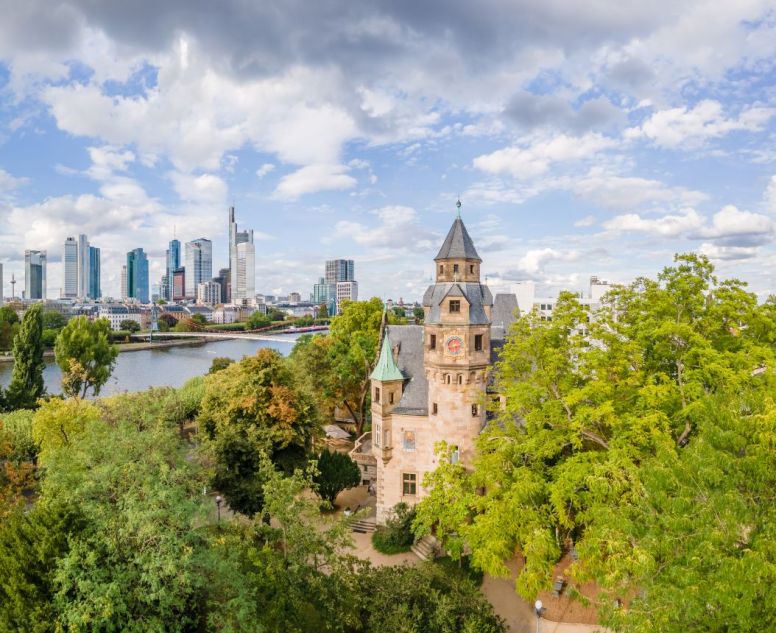 Alte Villa mit T&uuml;rmchen im Vordergrund in mitten von B&auml;umen, dahinter ein Fluss und Skyline mit Hochh&auml;usern auf der anderen Uferseite