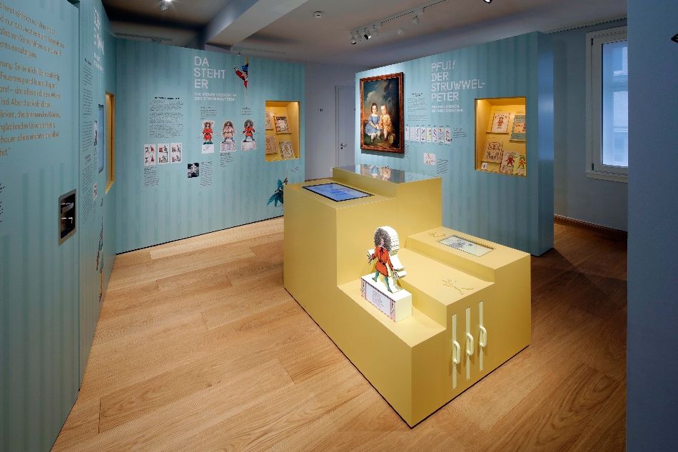 Ausstellung im Struwwelpeter Museum mit hellblauen Wänden und gelbem Erkundungstisch im Vordergrund