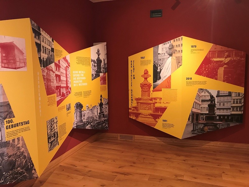 Gelbe Ausstellungswände mit Fotos und Erklärungstexten vor rotem Hintergrund