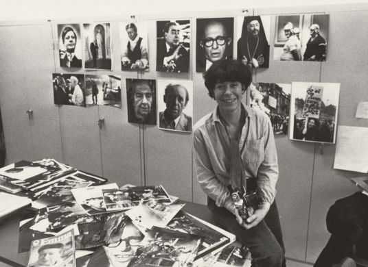 Schwarz-weiß Fotografie, auf der eine Frau mit kurzen dunklen Haaren auf einem Tisch sitzt mit vielen Fotos und Magazinen, im Hintergrund Portäts an der Wand