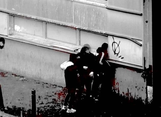 Verschwommenes schwarz-weiß Foto dreier Personen mit Jogginganzügen und Kopfbedeckung von hinten, die durch einen Fensterspalt in ein heruntergekommenes Gebäude blicken