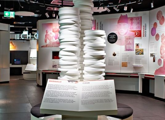 Leuchtschrift "Bargeld" mit großem, weißen Münzturm darunter im Vordergrund, dahinter Wand mit Texten und Bildern