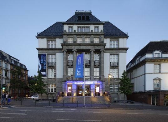 Fassade des Filmmuseum mit Straße im Vordergrund und blau erleuchtetem Eingangsbereich
