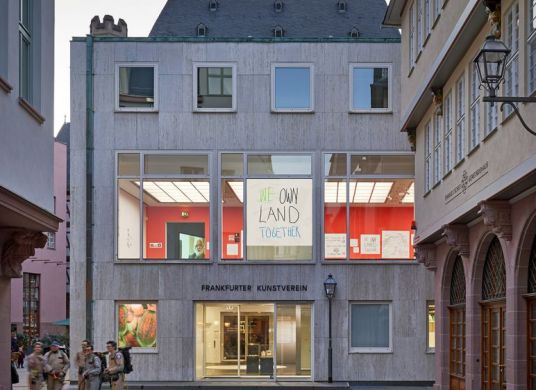 Gebäudeansicht des Frankfurter Kunstvereins, durch die Fenster sind Teile der Ausstellung zu sehen und ein großes Plakat auf dem in bunter Kreide steht "WE OWN LAND TOGETHER"