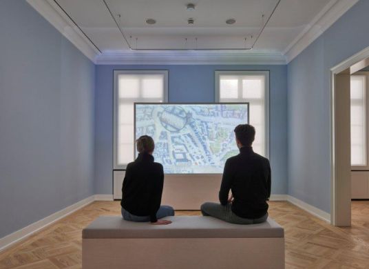 Zwei Menschen sitzen vor einer Medientafel in einem Raum