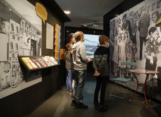 Mehrere Kinder schauen in einem Ausstellungsraum auf ein großes Schwarz-Weiß-Foto an der Wand auf dem Kinder und Männer in schwarzen Uniformen auf der Straße stehen