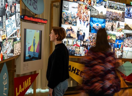 Zwei Personen stehen in Raum mit Bildschirmen und Nachrichtenbilderflut an den W&auml;nden und schauen diese an