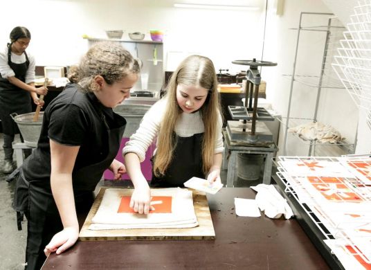 Zwei Mädchen beugen sich über einen Tisch in der Druckwerkstatt, wo sie einen Druck des Logos des Museums herstellen, im Hintergrund rührt ein weiteres Mädchen in einem Zuber