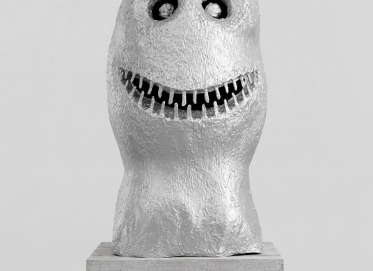 Eine Skulptur aus Aluminium, die wie ein vereinfachter grinsender Kopf aussieht, auf einem Betonsockel