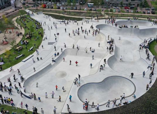 Gro&szlig;er Skatepark aus Beton mit verschiedenen Rampen von oben fotografiert mit vielen Menschen, drumherum Wiesen