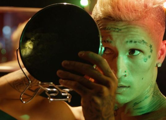 Eine Person mit kurzen Haaren und Tatoos im Gesicht schaut sich in einem Handspiegel an