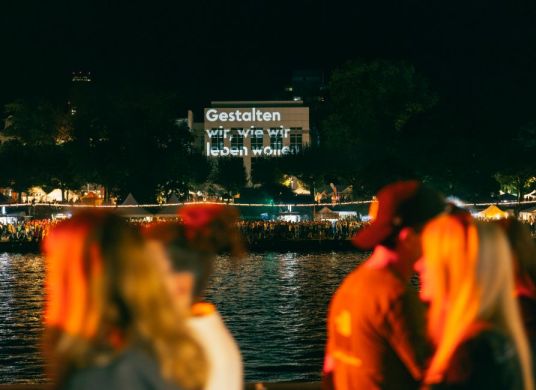 Menschen spazieren bei Nacht am Flussufer, auf ein Haus ist 'Gestalten wir, wie wir leben wollen' projiziert