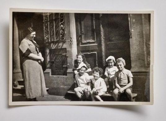  Auf einer alten Schwarz-Weiß-Fotografie schaut eine Frau auf fünf Kinder, die auf Treppenstufen vor einer Tür sitzen 