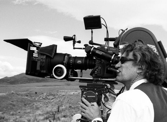 Schwarz-Weiß-Fotografie eines Mannes mit Sonnenbrille, der in einer kargen Landschaft steht und durch eine Filmkamera schaut