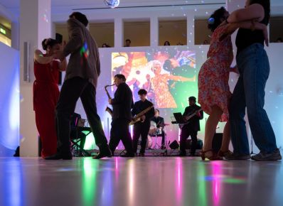 Zwei Paare tanzen in einem wei&szlig;en Raum vor einer Band und einer Projektion an der Wand, durch Durchbr&uuml;che in der Wand schauen von oben Personen zu