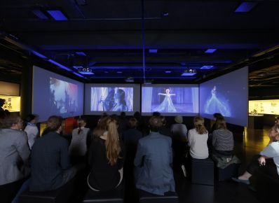 Blick in den abgedunkelten Filmraum im DFF - Deutsches Filminstitut & Filmmuseum mit mehreren Bildschirmen und Sitzgelegenheiten und Besucher*innen