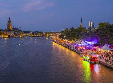Erleuchtetes Ufer eines Flusses mit Buden und vielen Menschen von Weitem mit Brücke, Hochhaus und Kirchen im Hintergrund